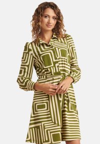 Groen en crèmekleurige jurk met patroon, lange mouwen, knoopsluiting aan de voorkant en tailleband. Bevat geometrische vormen en heeft een zachte, gladde textuur.