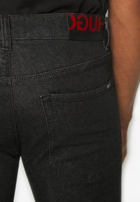 Vaqueros de mezclilla negros con una cinturilla de logo roja, que cuentan con un bolsillo trasero y una textura suave. Diseño sencillo con costuras en contraste.