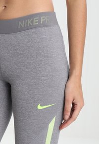 Persona vistiendo leggings grises de Nike Pro con el logo en neón amarillo y detalles de diseño a juego en el muslo, con la mano descansando a un lado.