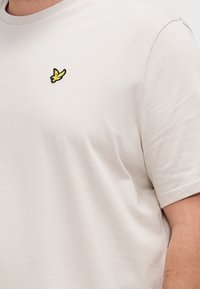 T-shirt beige clair en coton, avec un col rond. Il présente un petit logo brodé de banane noir et jaune sur la poitrine.