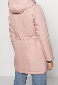 Ljusrosa vadderad jacka med huva, bälte i midjan och elastiska ärmslut. Slät yta med sömdetaljer längs fållen och sömmarna.