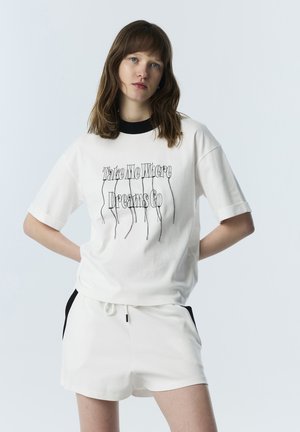 Jonge vrouw met bruin haar draagt witte shorts en een wit T-shirt met zwarte kraag en tekst "Neem me mee waar dromen naartoe gaan."