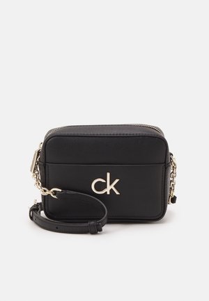 Black rectangular leather crossbody bag with gold "ck" logo, chain-link strap, and zipper closure, shown on a white background.