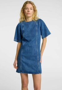 Denimkjole i medium blå med korte ermer, rund hals og synlige sømmer. Stoffet har en glatt tekstur og en lett tettsittende fasong.