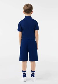 Bambino in piedi di spalle che indossa una polo blu navy, pantaloni corti abbinati, calzini blu con strisce bianche e sneaker bianche su uno sfondo bianco.