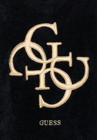 Črn material z zlatim vezenim dvojnim logotipom 'G', okrašenim s krivuljami in bisernim obrobjem. Ime blagovne znamke "GUESS" je spodaj.
