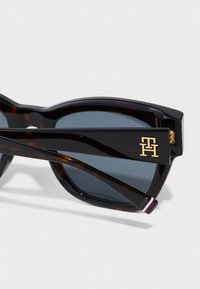 Ochelari de soare cu model țestoasă, cu lentile gri închis. Prezintă un logo aurit pe braț, un design unic al ramei și un accent în dungi pe temple.