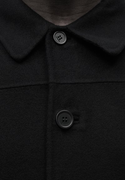 Μαύρο μάλλινο παλτό με δύο μεγάλα μαύρα κουμπιά και φαρδύ γιακά, εμφανίζοντας μια κοντινή εικόνα του επάνω μπροστινού μέρους.