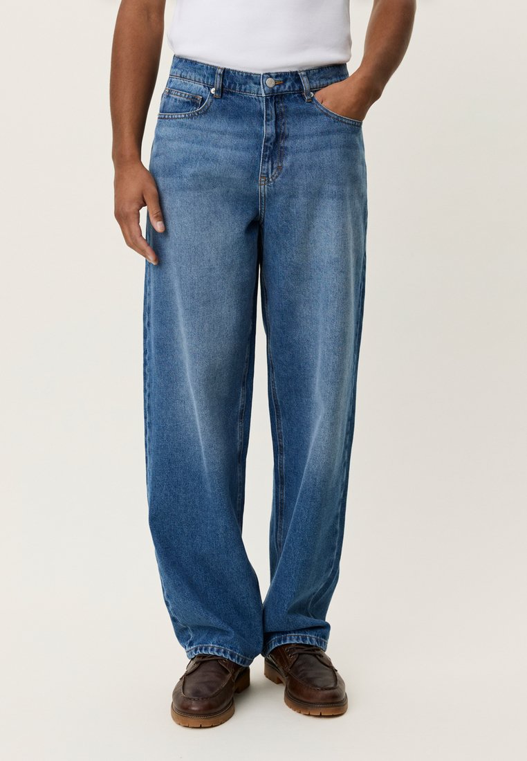 Μπλε τζιν παντελόνι με χαλαρή εφαρμογή, διαθέτει μπροστινή τσέπη, θηλιές για ζώνη και διακριτική ξεθώριαση. Συνοδεύεται από καφέ υποδήματα.