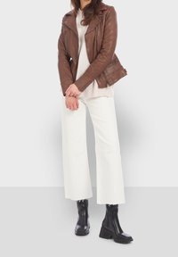 Brun läderbikerjacka över en krämfärgad tröja med vita vida byxor och svarta chunky klackstövlar, som visar dragkedjor och strukturerade detaljer.