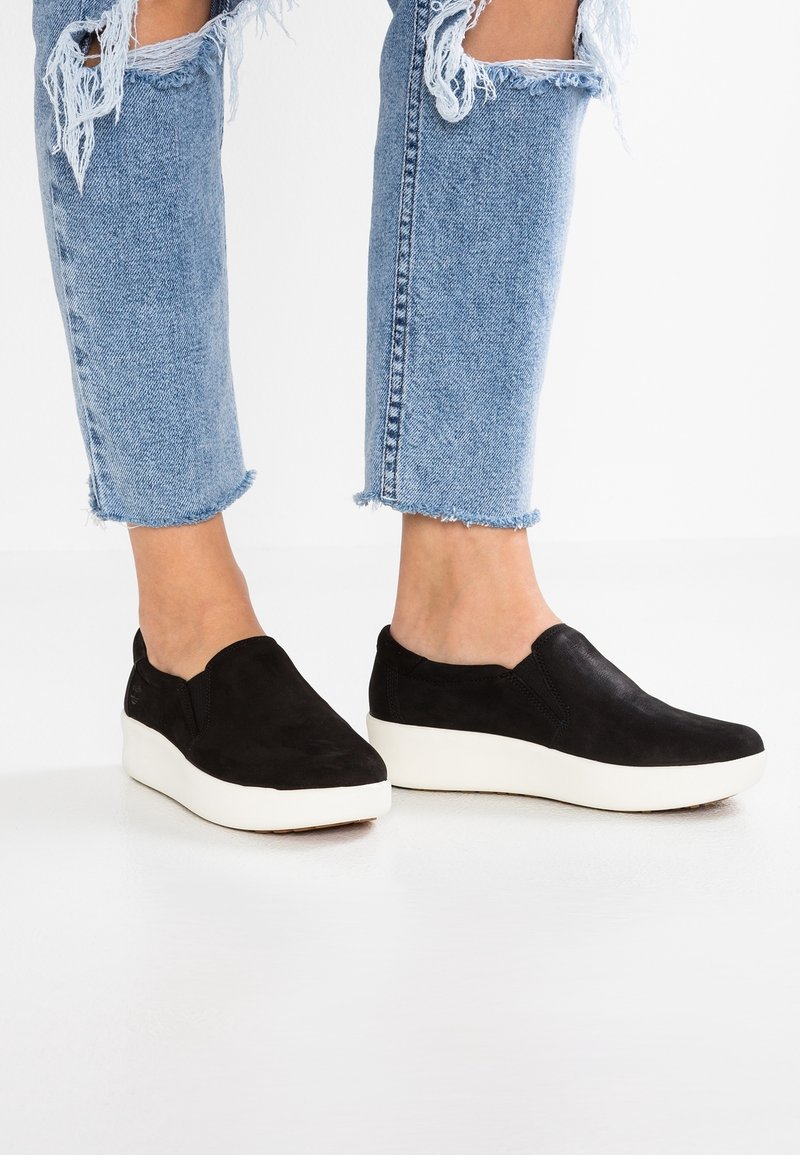 Personne portant des baskets slip-on noires avec des semelles blanches épaisses et un jean bleu clair déchiré, coupé au-dessus de la cheville.
