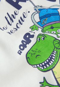 Dinosaure vert dessin animé et jouet extraterrestre à trois yeux avec tenue bleue, accompagné du texte "to the rescue" et "ROAR" sur fond blanc.