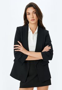 Czarna marynarka z wyraźnymi ramionami, dwiema przednimi kieszeniami i lekko oversize'owym krojem, zestawiona z białą bluzką z kołnierzykiem i czarną spódnicą.