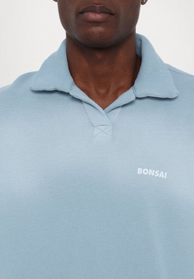 Vaaleansininen polo-paita, jossa on pehmeä texture, klassinen kaulus ja pieni valkoinen "BONSAI" -logo rinnassa.