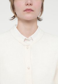 Cardigan de cor creme com textura canelada e decote redondo, apresentando quatro pequenos botões de pérola na frente, usado sobre uma camisa branca com colarinho.