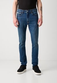 Schmal geschnittene blaue Jeans mit Fünf-Taschen-Design, leichter Ausbleichung und mittelhohem Bund. Kombiniert mit schwarzen Sneakers.