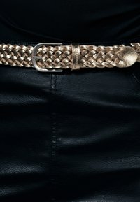 Cintura in pelle dorata intrecciata con una fibbia metallica argentata, posizionata su un capo d'abbigliamento in pelle nera, che mette in risalto la texture e il design intricato.