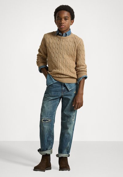 Bežs adīts džemperis virs zila džinsa krekla, kombinēts ar nolietotiem ziliem džinsiem un brūnām potīšu zābakiem. Vienkāršs, ikdienišķs dizains.