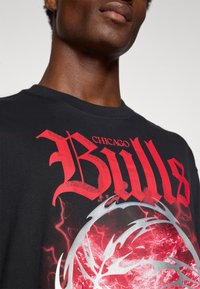 T-shirt de algodão preta com grande texto em vermelho "Bulls" e elementos gráficos prateados em espiral, criando um design dinâmico.