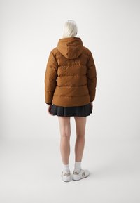 Brown puffer jacket with a hood, quilted design, and elastic cuffs. Worn over a black skirt paired with white sneakers and crew socks.