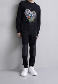 Felpa nera con una grafica di un cuore a strisce zebrate e un coniglio dei cartoni animati. Pantaloni neri con toppe nere a contrasto. Sneakers bianche. Texture liscia.