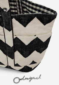 Black and cream zigzag patterned tote bag made of canvas, featuring two exterior pockets and a silver logo accent.
