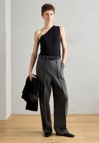 Top negro de un solo hombro, pantalones de talle alto grises con pliegues, cinturón negro y sosteniendo un bolso negro texturizado. Fondo de suelo de madera.