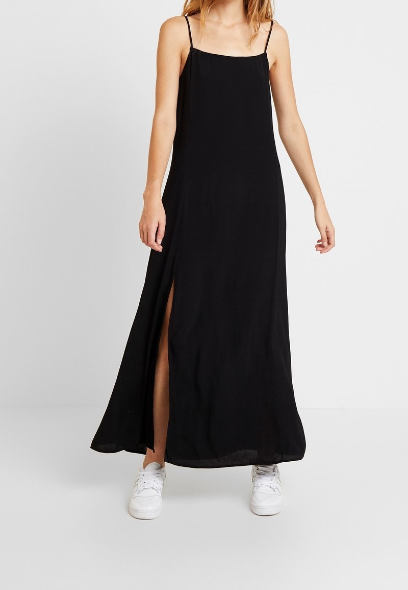 Femme portant une robe maxi noire sans manches avec une fente latérale et des baskets blanches, debout devant un fond clair uni.