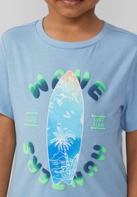 Hellblaues Baumwoll-T-Shirt mit einem Grafikdesign eines Surfbretts, umgeben von Palmen, mit grünem und dunkelblauem Text "WAVE SURF TEAM".