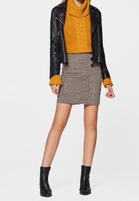 Chaqueta de cuero negro de motorista sobre un suéter de punto en color mostaza, con una falda ajustada de cuadro de gallo y botas negras hasta el tobillo.