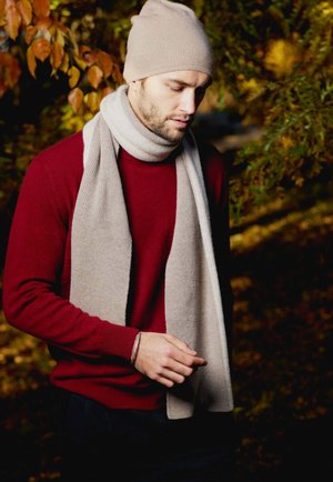 Berretto a maglia beige e sciarpa a coste grigio chiaro indossati sopra un maglione rosso scuro. L'uomo ha una leggera barba ed è in un ambiente naturale all'aperto.