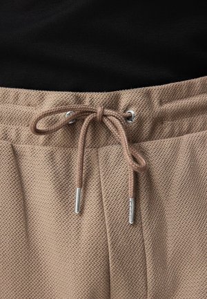 Lichtbruine shorts met textuur en een elastische tailleband, voorzien van een trekkoord met zilveren punten. Zwarte top gedeeltelijk zichtbaar bovenaan.