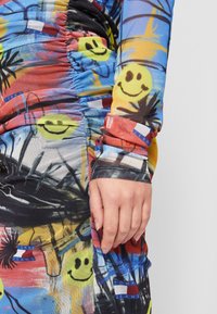 Mano descansando sobre tela colorida con pliegues que presenta caritas sonrientes, formas abstractas en negro y logotipos de Tommy Jeans en rojo, azul, amarillo y blanco.