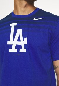 Camiseta de manga corta azul, hecha de un tejido texturizado, que presenta un gran logo blanco de "LA" y un pequeño swoosh blanco de Nike en la parte superior del pecho.