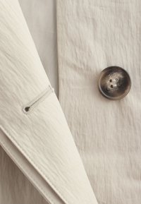 Gros plan d'un tissu beige avec un grand bouton marron et une manche repliée présentant une boutonnière.