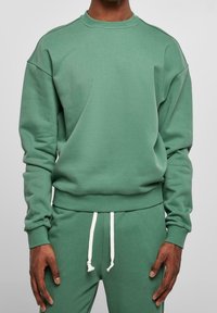 Sweat-shirt vert à col rond côtelé et épaules tombantes, avec une texture lisse et des manches resserrées aux poignets, porté avec un pantalon de survêtement vert assorti.