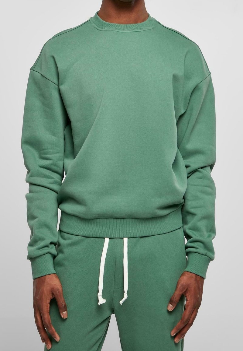 Sweat-shirt vert à col rond côtelé et épaules tombantes, avec une texture lisse et des manches resserrées aux poignets, porté avec un pantalon de survêtement vert assorti.