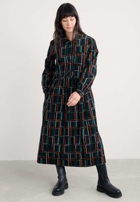 Crna baršunasta haljina s kariranim uzorkom u šarenim linijama. Ima ovratnik, duge rukave i cinčanu struk. Manekenka nosi crne čizme.