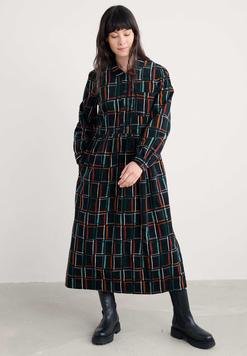 Crna baršunasta haljina s kariranim uzorkom u šarenim linijama. Ima ovratnik, duge rukave i cinčanu struk. Manekenka nosi crne čizme.