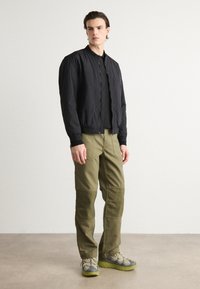Chaqueta bomber negra con tejido texturizado, combinada con pantalones cargo verde oliva con detalles de patchwork y zapatillas gris-verde.