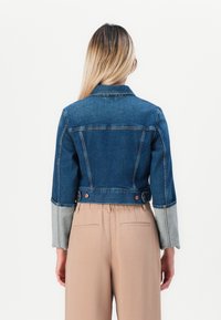 Woman with long blonde hair wears cropped two-tone denim jacket and beige high-waist pants, standing against plain white background.
