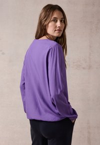 Blouse violette à manches longues avec une coupe décontractée, texture douce et encolure ronde ; portée avec un pantalon noir, présentée sur fond neutre.