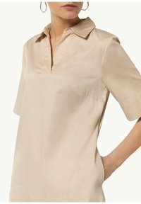 Blusa beige de manga corta con cuello y escote en V. Confeccionada en tejido suave, presenta un ligero brillo y un bolsillo lateral.