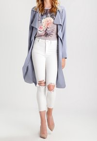 Lysegrå lang frakke over en blomstret top, kombineret med hvide slidte skinny jeans og nøgenfarvede høje hæle. Glat stof med blød fald.