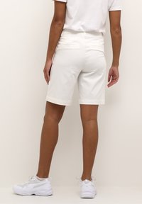 Shorts bianchi in cotone con orli risvoltati, due tasche posteriori e una vestibilità a vita media, abbinati a scarpe da ginnastica bianche.