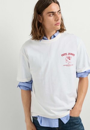 Giovane uomo con capelli alla lunghezza delle spalle che indossa una maglietta bianca Pepe Jeans sopra una camicia blu con bottoni, guardando di lato.