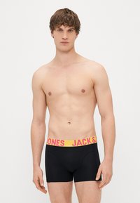 Černé boxerky s širokým žlutým páskem a růžovým nápisem. Model má neutrální výraz a je bez trička.