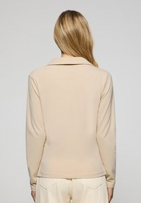 Beige långärmad skjorta med krage, med en slät textur och en något åtsittande design, visad från baksidan.