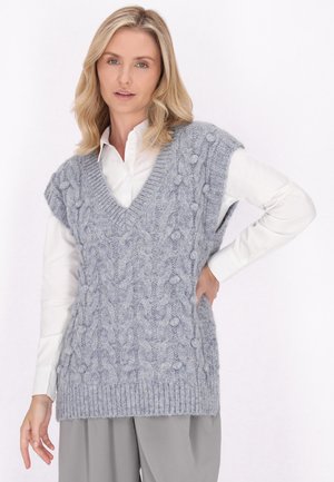 Lichtgrijze gebreide vest met een V-hals, kabelpatroon en getextureerde bobbels. Draagt over een wit shirt en gestyled met grijze broek.