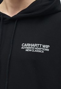 Musta huppari, jossa on valkoinen teksti "CARHARTT WIP AUTHENTIC ADAPTIONS NEW CLASSICS" painettuna rinnan kohdalle, lähikuva ylävartalosta.